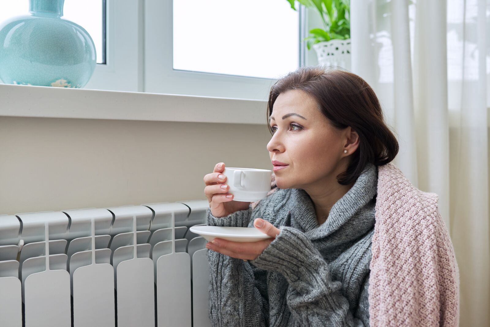 Frau an Heizkörper drinnen schaut durch Fenster und trinkt Tee