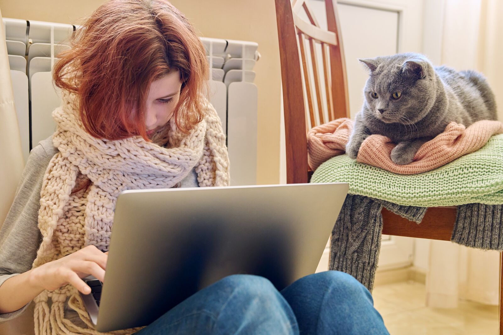 Eine Frau mit einem dicken Schal und einem Rechner auf den Knien. Eine Katze schaut ihr zu.