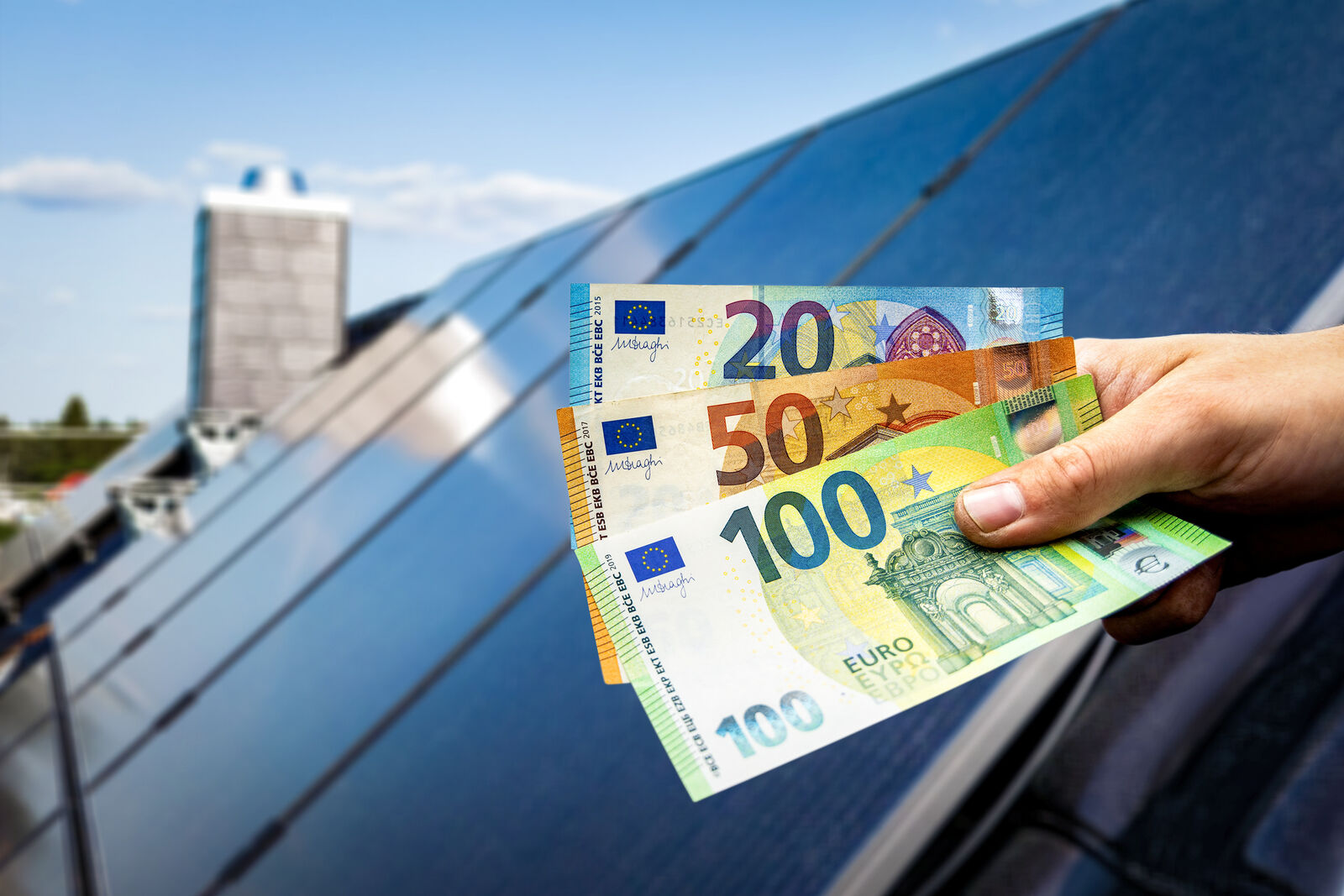 Eine Hand hält drei Euroscheine vor einem Dach mit Solarpanelen darauf.