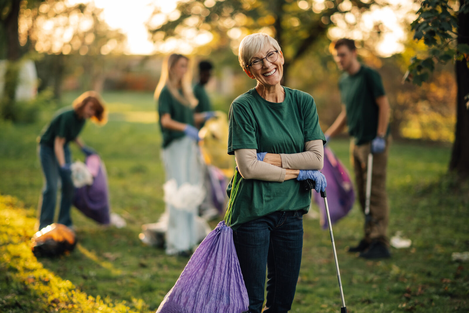 eine Gruppe von Menschen on grünen T-Shirts sammelt Müll in Beutel, eine kurzhaarige ältere Frau mir Brille schaut dabei direkt in die Kamera