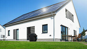 Ein weißes Haus mit Solarpanels auf dem Dach vor blauem Himmel, vor dem Haus ist ein grüner Rasen