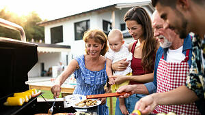Großfamilie am Grill mit Hintergrund Haus, darauf liegt Fleisch und Gemüse