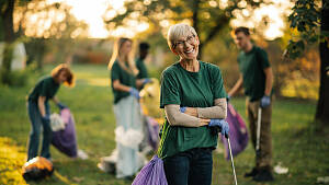 eine Gruppe von Menschen on grünen T-Shirts sammelt Müll in Beutel, eine kurzhaarige ältere Frau mir Brille schaut dabei direkt in die Kamera