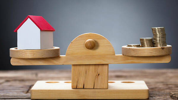 Themenbild: eine Waage aus Holz, in der einen Waggschale ein Hausmodell, in der anderen einige Münzstapel