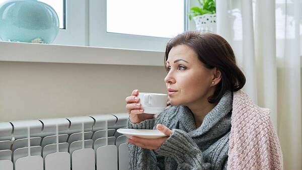 Themenbild: Frau an Heizkörper drinnen schaut durch Fenster und trinkt Tee
