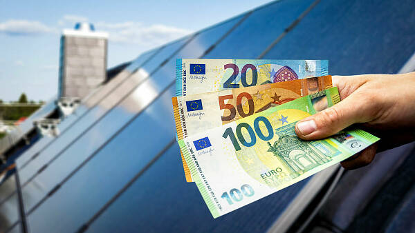 Themenbild: Eine Hand hält drei Euroscheine vor einem Dach mit Solarpanelen darauf. Themenbild: Eine Hand hält drei Euroscheine vor einem Dach mit Solarpanelen darauf.