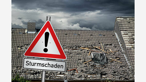 Themenbild: Ein Warnschild mit der Aufschrift Sturmschäden vor einem Haus mit einem kaputten Dach